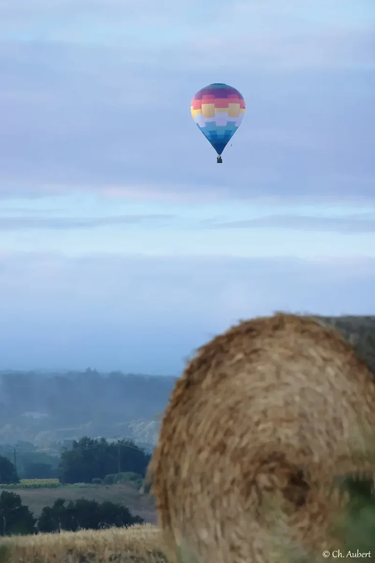 Montgolfière multicolore de L'Air du Perche flottant au loin, avec une botte de foin au premier plan.