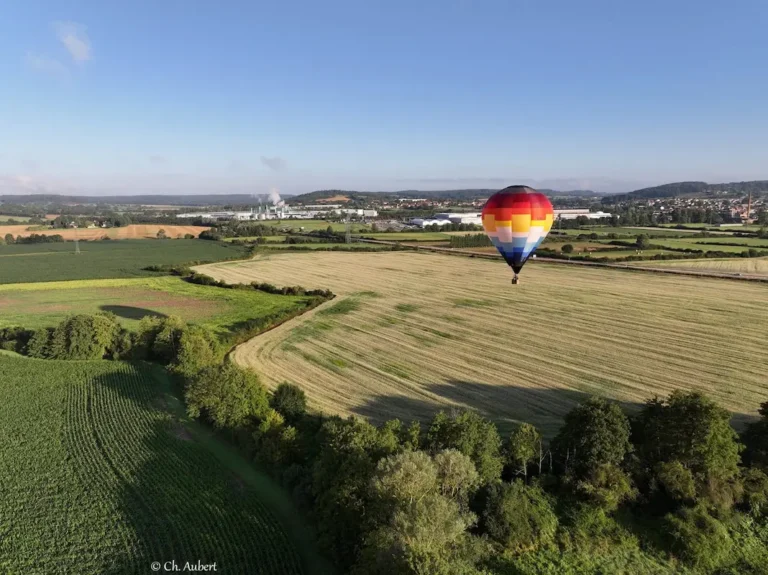 Vue aérienne de la montgolfière de L'Air du Perche survolant des champs avec un village en arrière-plan.